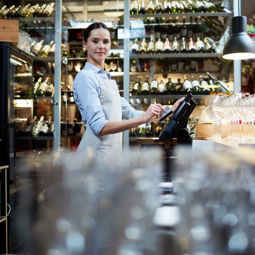Young staff of restaurant standing by computer and entering data about latest orders and payments