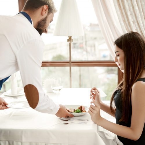 Handsome waiter at the restaurant is serving a young lady a meal